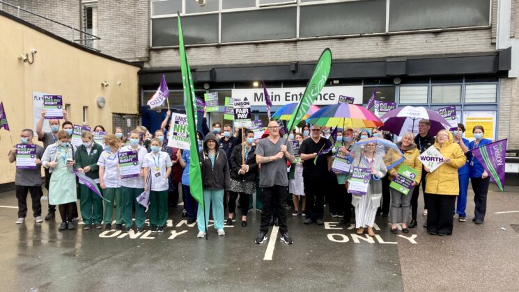 Health care staff brave the rain with umbrellas outside Derriford Hospital. They hold Put NHS Pay Right signs.