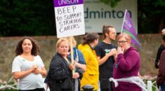 Care workers taking part in strike action picket outside a workplace. One carer holds a UNISON placard which says 'Beep to support striking carers'