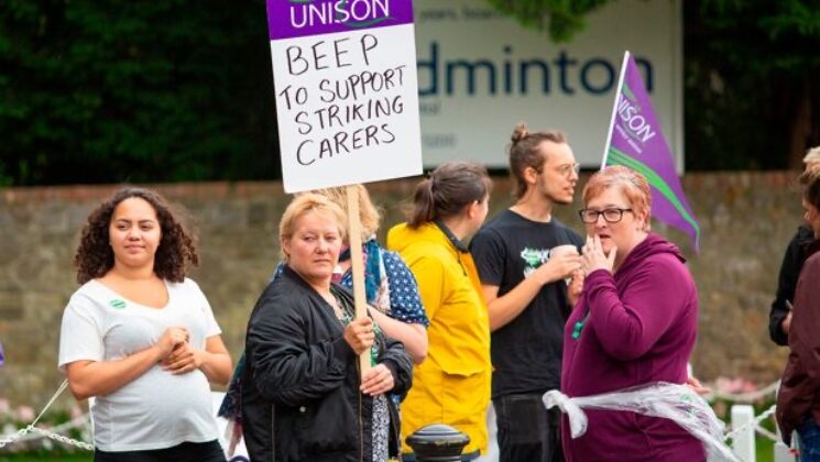Care workers taking part in strike action picket outside a workplace. One carer holds a UNISON placard which says 'Beep to support striking carers'