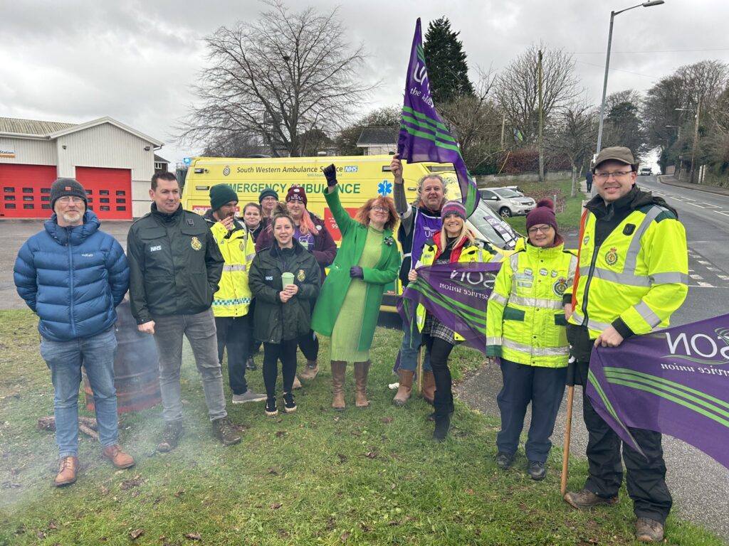 Joanne Kay raises a fist alongside picketing ambulance staff. An ambulance can be seen in the background and smoke rises from the nearby brazier.