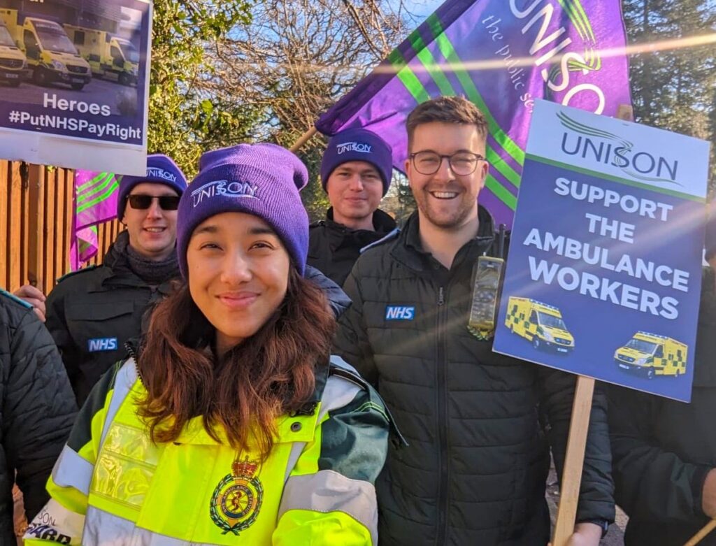Alice Foxen on the picket line with ambulance workers, they hold UNISON flags and placards.