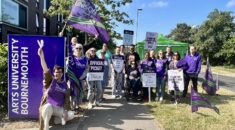 UNISON members on the picket line outside Arts University Bournemouth. Members hold purple flags and placards aloft against a beautiful blue sky.