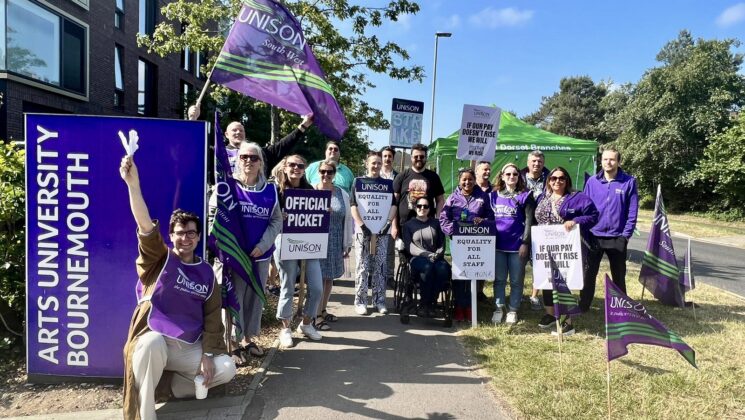 UNISON members on the picket line outside Arts University Bournemouth. Members hold purple flags and placards aloft against a beautiful blue sky.