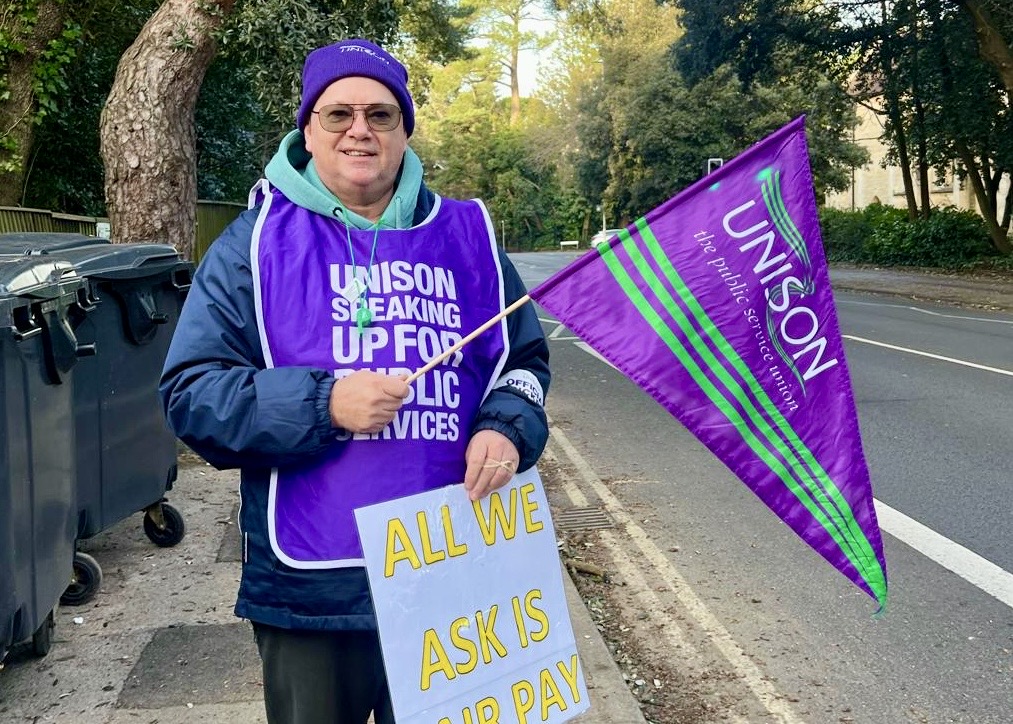 UNISON member in purple tabard holding a UNISON flag on the picket line in Dorset.