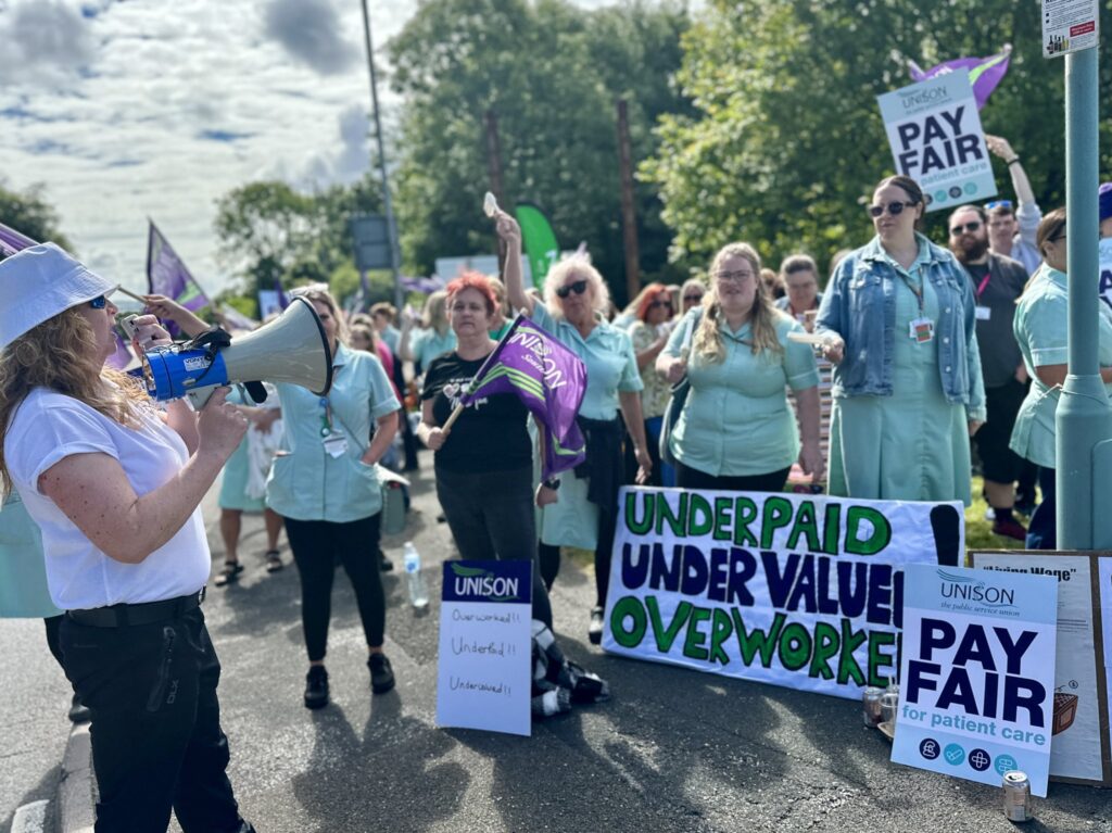 Karen holds a megaphone as healthcare workers stand behind a handmade sign which reads “underpaid, undervalued, overworked”