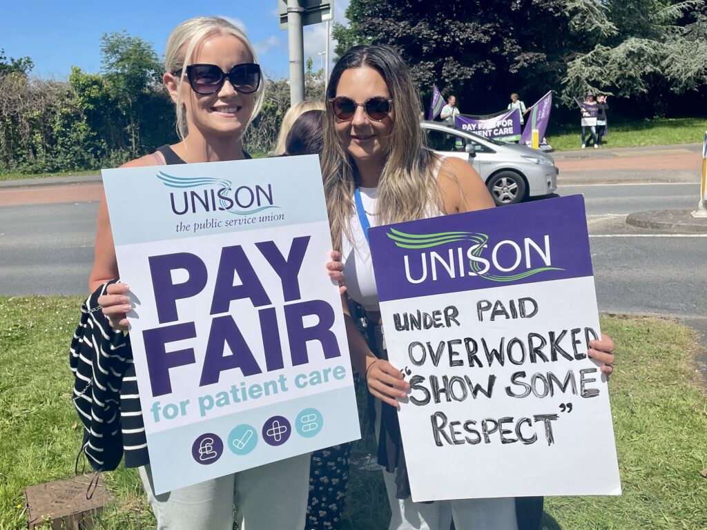 Two healthcare workers on the picket line hold placards. One reads “pay fair for patient care” and the other says “underpaid, overworked, show some respect”
